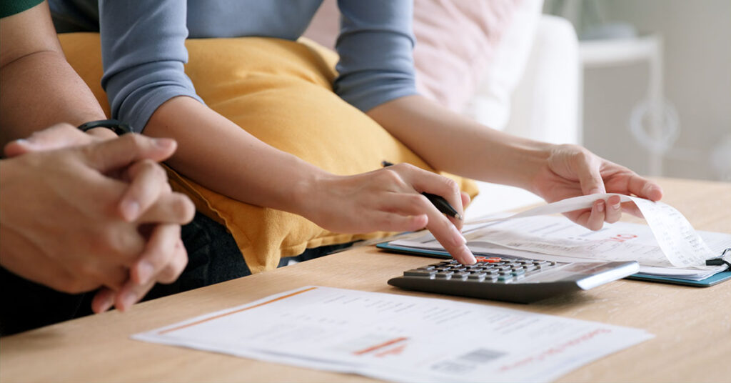 A close-up of two people reviewing financial documents at home. One person is using a calculator while holding a receipt, and various papers with financial information are spread on the table.