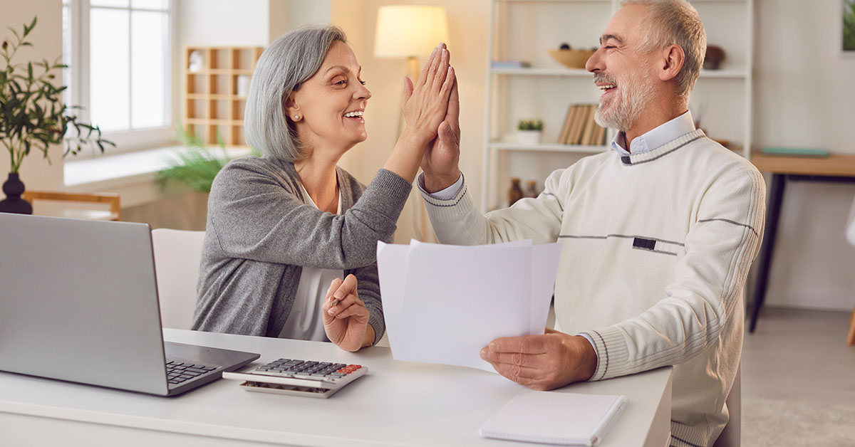 Senior couple celebrating financial success while working on tax planning with a laptop, calculator, and documents in a well-lit home office.