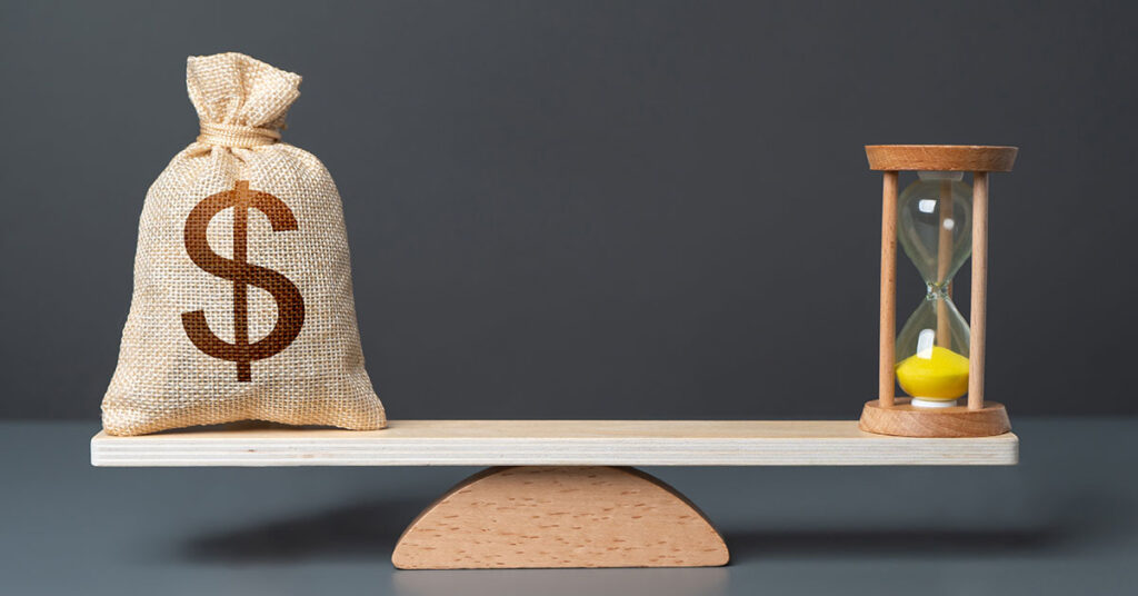 A burlap sack with a dollar sign and an hourglass sit on opposite ends of a wooden balance, symbolizing the tradeoff between money and time in investment decisions.