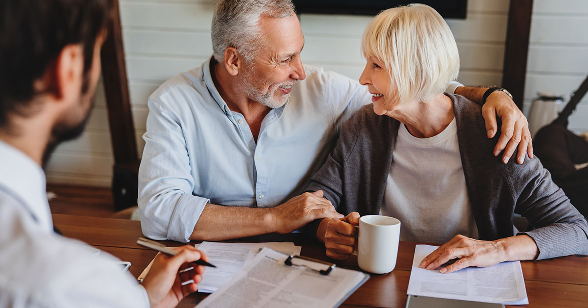 Smiling older couple sit together at a table, holding hands and a mug, speaking with a financial advisor (seen from behind); documents and a clipboard are on the table.