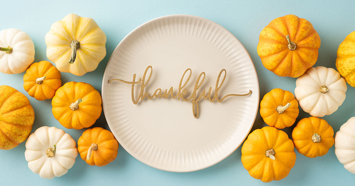 A white plate with the word "thankful" in golden cursive, surrounded by orange and white pumpkins on a blue background, symbolizing gratitude and abundance—key themes explored in the article about thankfulness and financial well-being.