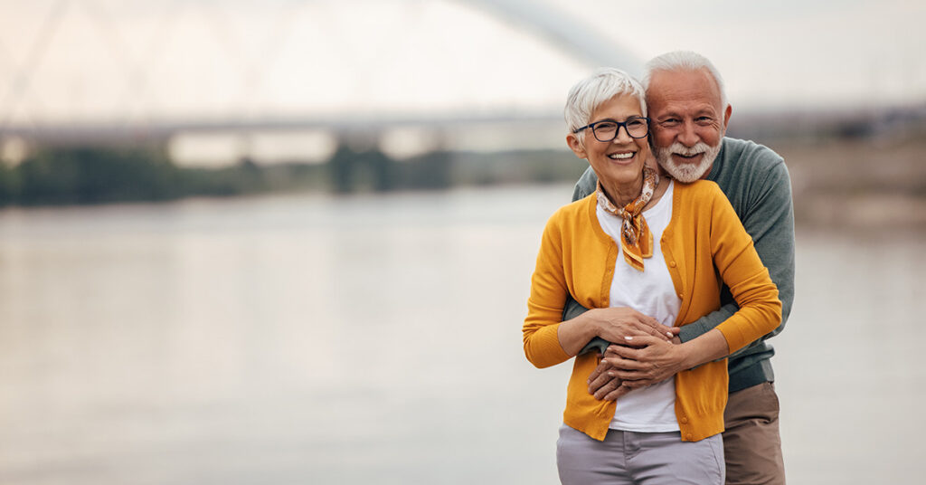Smiling older couple standing by a river, with the man hugging the woman from behind as they look at the camera.