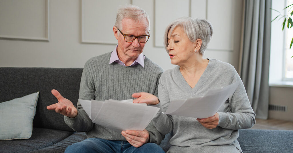 An older couple sitting on a couch reviewing paperwork together, looking concerned and discussing the documents in their hands.