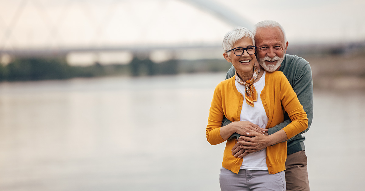 Smiling older couple standing by a river, with the man hugging the woman from behind as they look at the camera.