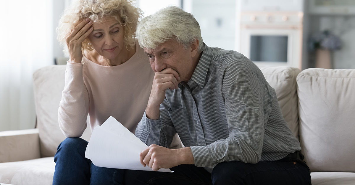 An older couple sits on a couch, reviewing financial documents with concerned expressions.