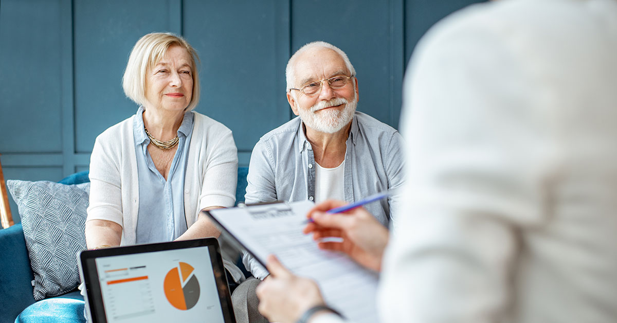 An older couple sits together and smiles while listening to a financial advisor during a meeting, with a laptop showing a pie chart in the foreground.