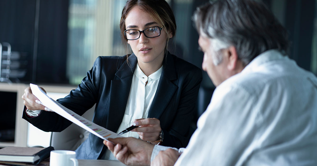Young female financial professional in glasses reviews a printed chart with an older male client at a desk.