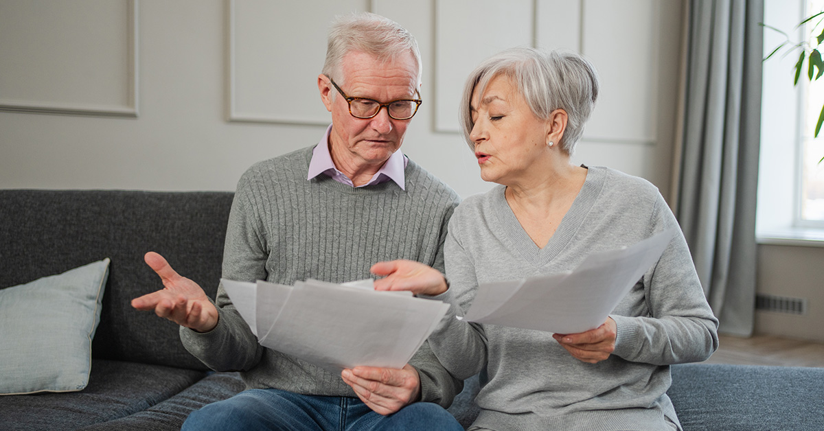 An older couple sitting on a couch reviewing paperwork together, looking concerned and discussing the documents in their hands.