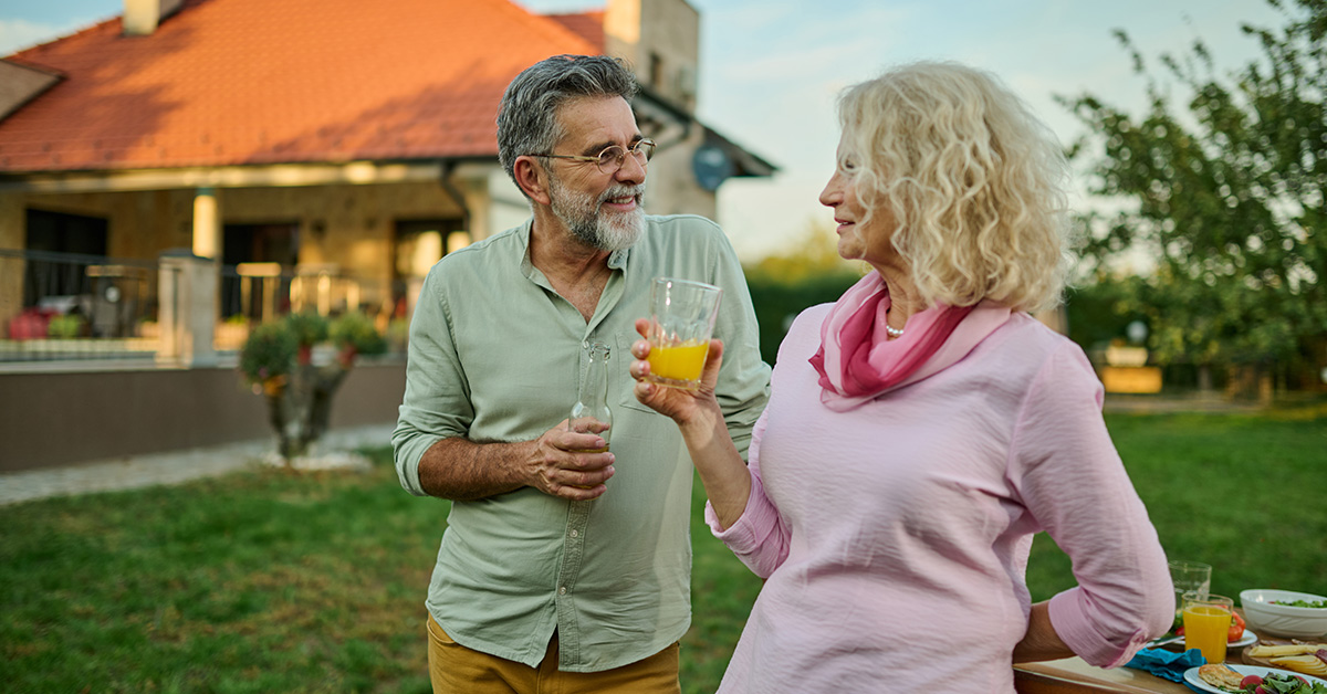 Active older couple standing in their backyard near a table of food, smiling at each other and holding drinks on a sunny day.